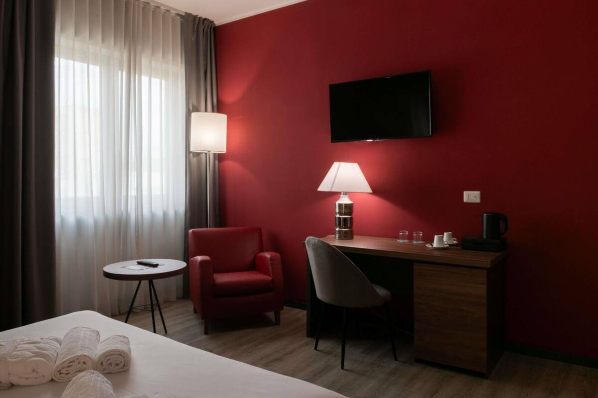 Habitación de hotel con cama, escritorio, silla, televisor, lámpara, cafetera y una mesita con toallas.