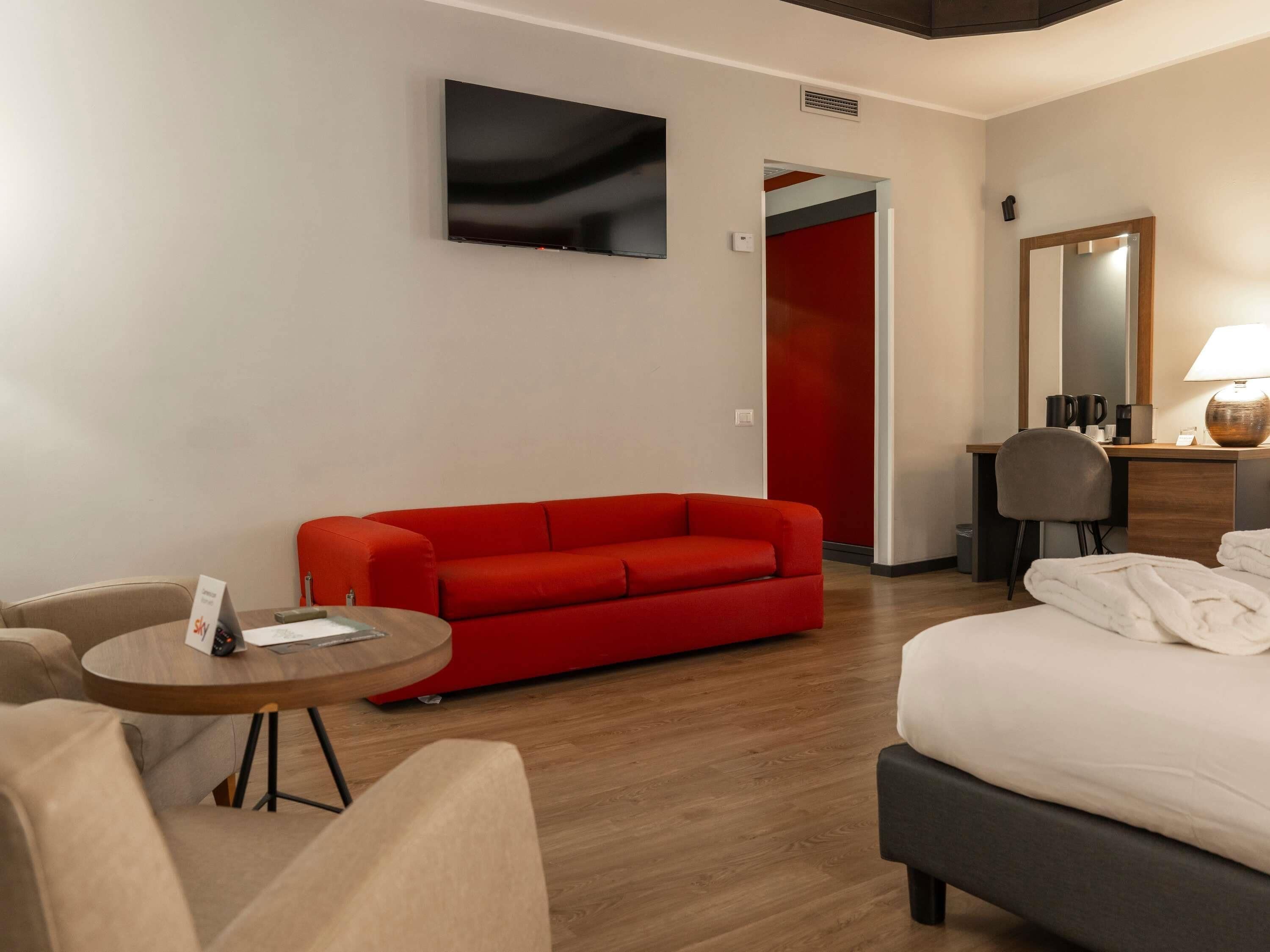 A hotel room with a red sofa, a round wooden table, a bed with white linens, a desk with a lamp, and a large mirror.