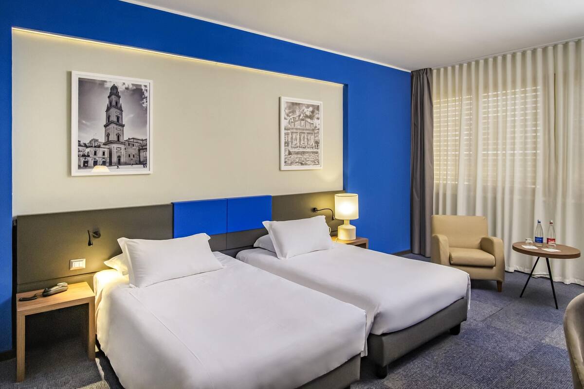 A hotel room with two beds, a chair, a small table, and two wall-mounted lamps.