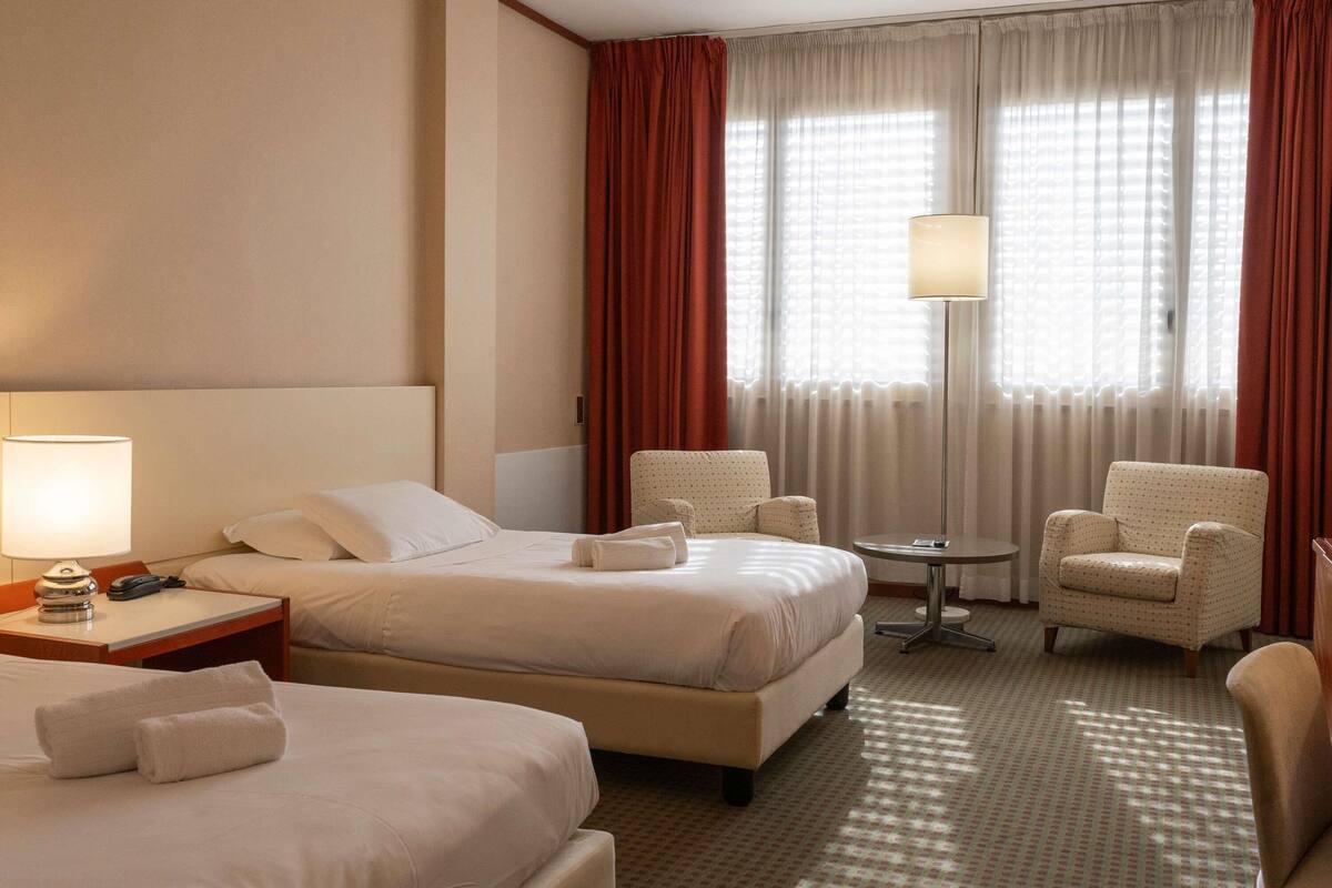 A hotel room with two beds, a desk with a lamp, a chair, and a small table.