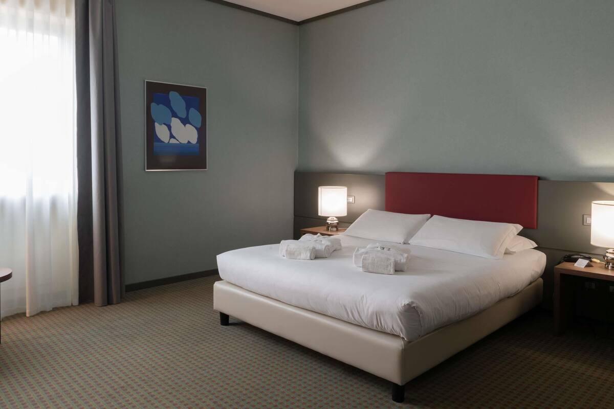 A hotel room with a bed, two bedside tables, a nightstand, a wall-mounted lamp, and a window with curtains.