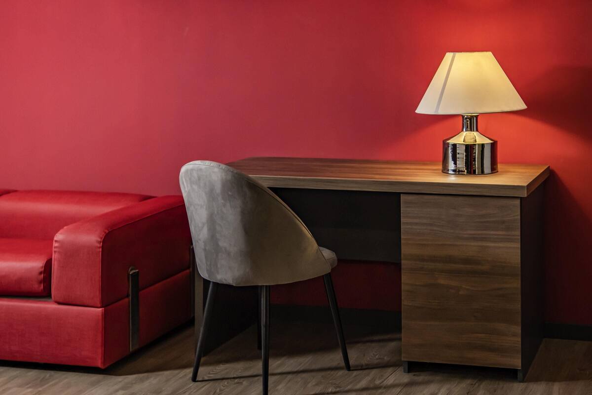 A red sofa, a wooden desk with a lamp, and a chair in a room with wooden flooring.