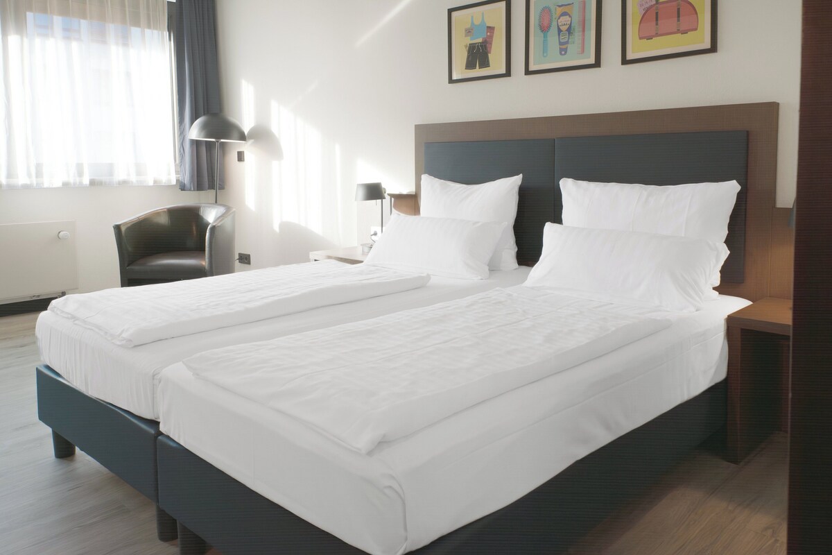 A hotel room with two beds, a desk, a chair, and a lamp.
