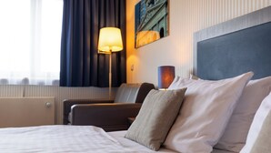 Habitación de hotel con cama, mesita de noche, lámpara, sillón y un cuadro en la pared.