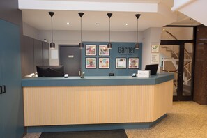 A hotel reception desk with a blue and beige color scheme, multiple pendant lights, framed artwork on the wall, and a staircase in the background.
