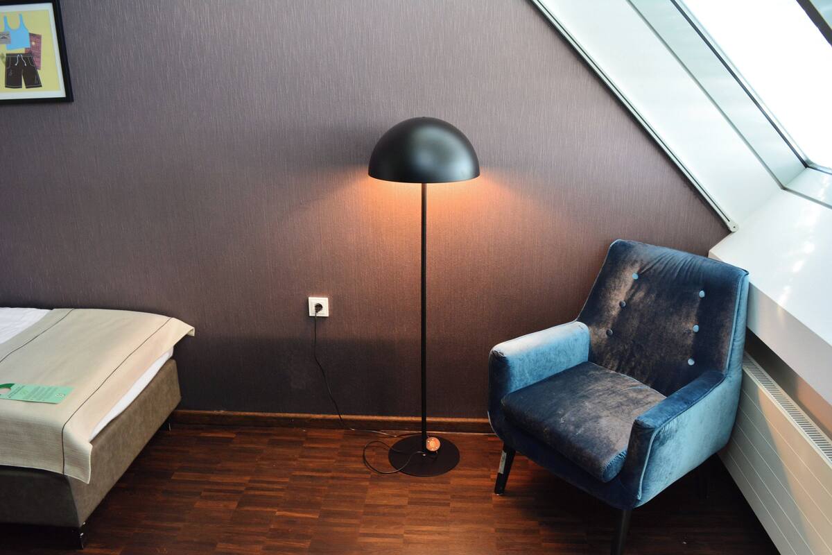 A room with a bed, a blue armchair, a floor lamp, and a window with a view of the sky.