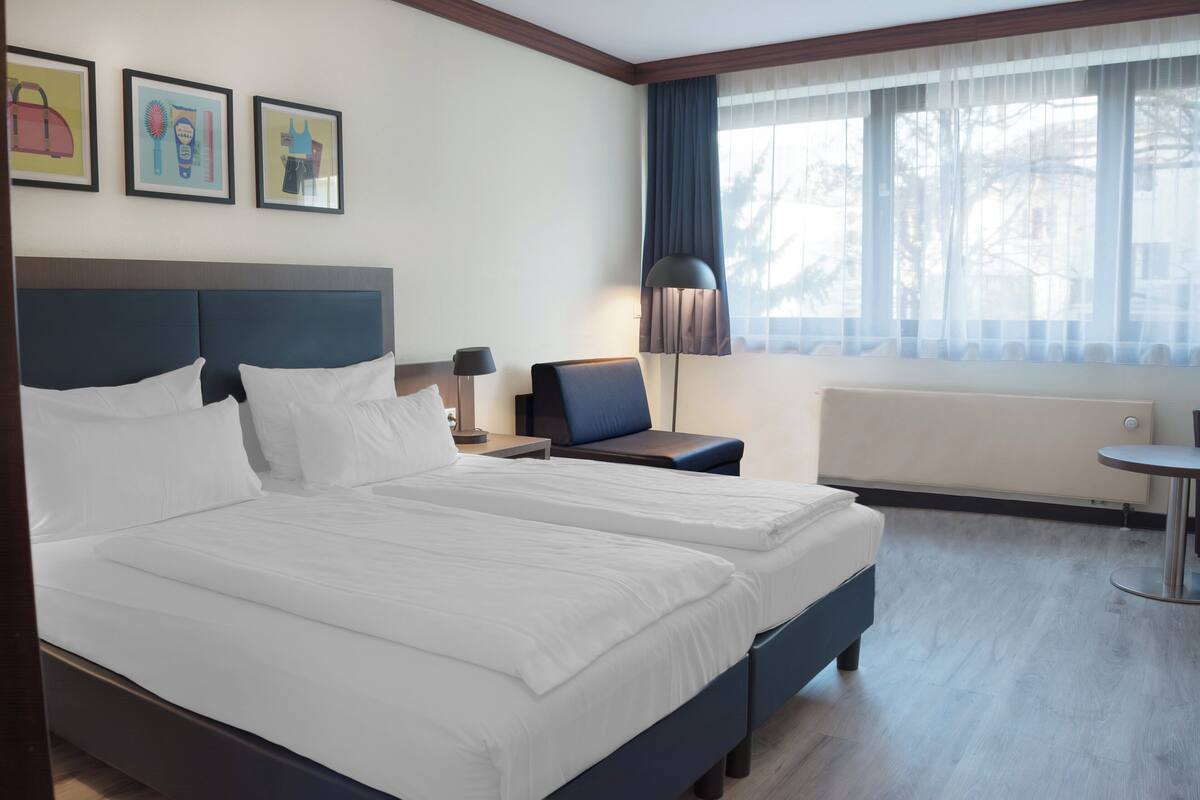 A hotel room with two beds, a desk, a chair, a lamp, and a window with curtains.
