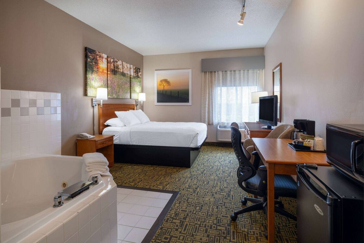A hotel room with a bed, a desk, a television, a refrigerator, a bathroom with a bathtub, and a large window.