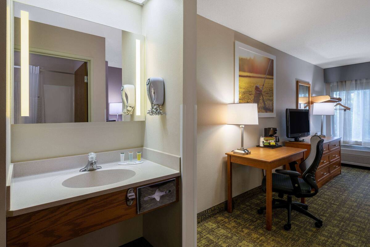 A hotel room with a bathroom featuring a sink, mirror, and towel rack. The room includes a desk with a chair, a television, and a window with curtains.