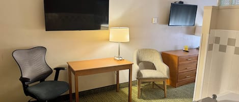 Una camera d'albergo con una scrivania, due sedie, una TV, una lampada, un settimino in legno e una vasca da bagno.