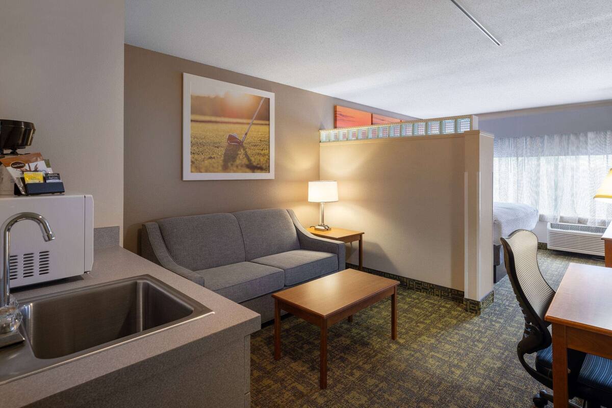 A compact hotel room with a kitchenette, a gray sofa, a wooden coffee table, a lamp, and a view of a field through the window.