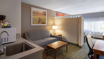 A compact hotel room with a kitchenette, a gray sofa, a wooden coffee table, a lamp, and a view of a field through the window.