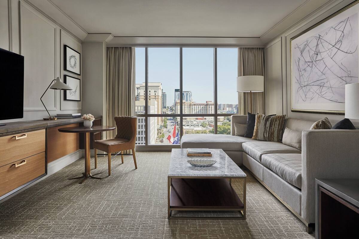 Ein modernes Hotelzimmer mit großem Fenster, einer Couch, einem Couchtisch, einem Fernseher und einem Schreibtisch.