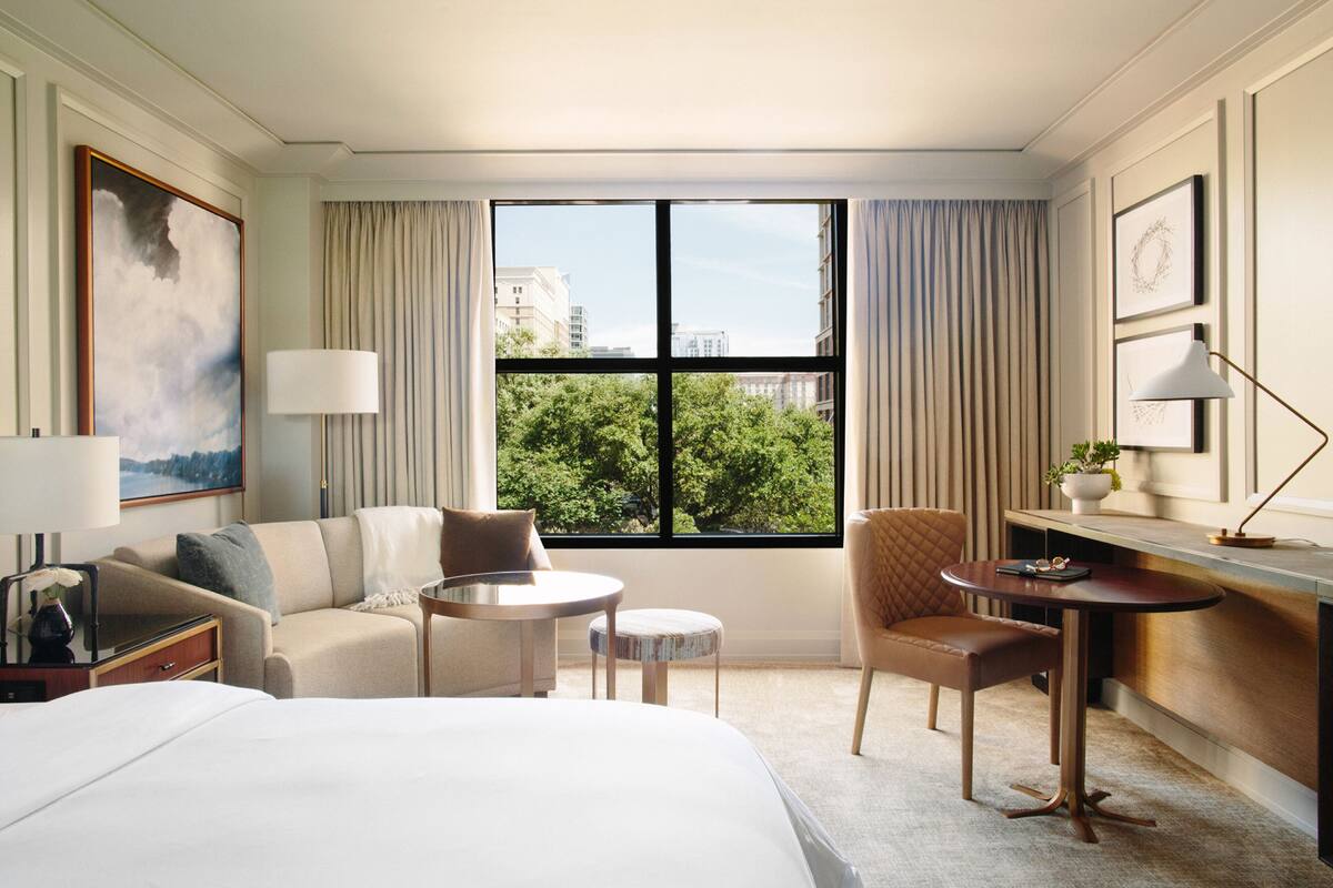 Ein Hotelzimmer mit Bett, einer Couch, einem runden Tisch, einem Sessel, einem Schreibtisch und Blick auf Bäume und Gebäude durch große Fenster.