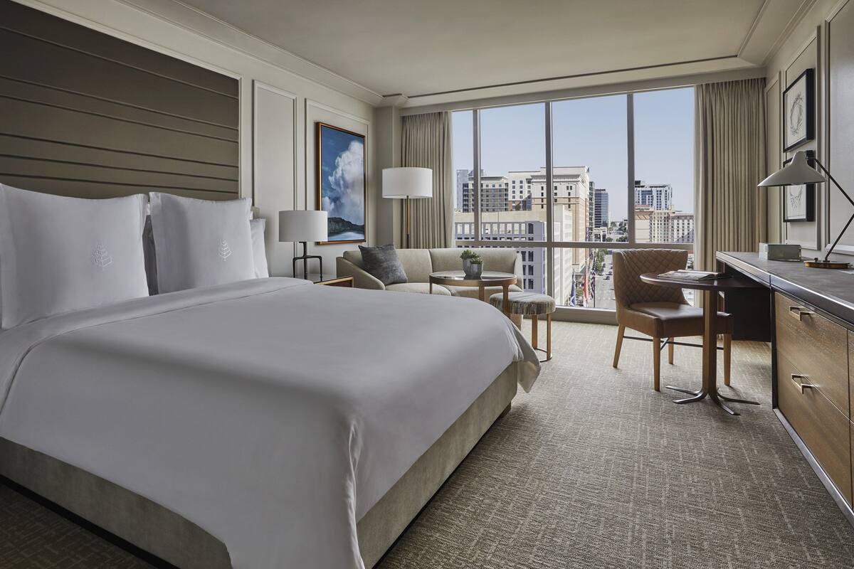 Ein Hotelzimmer mit einem großen Bett, einem Schreibtisch, einem Sessel und Blick auf die Stadt durch große Fenster.
