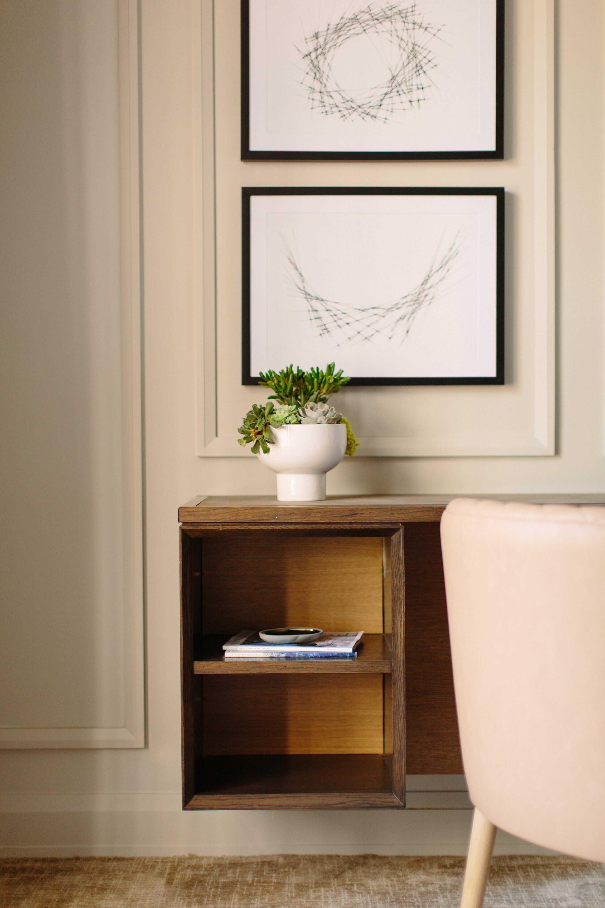 A framed abstract artwork on a wall, a white vase with a plant on a wooden shelf, and a light-colored chair.