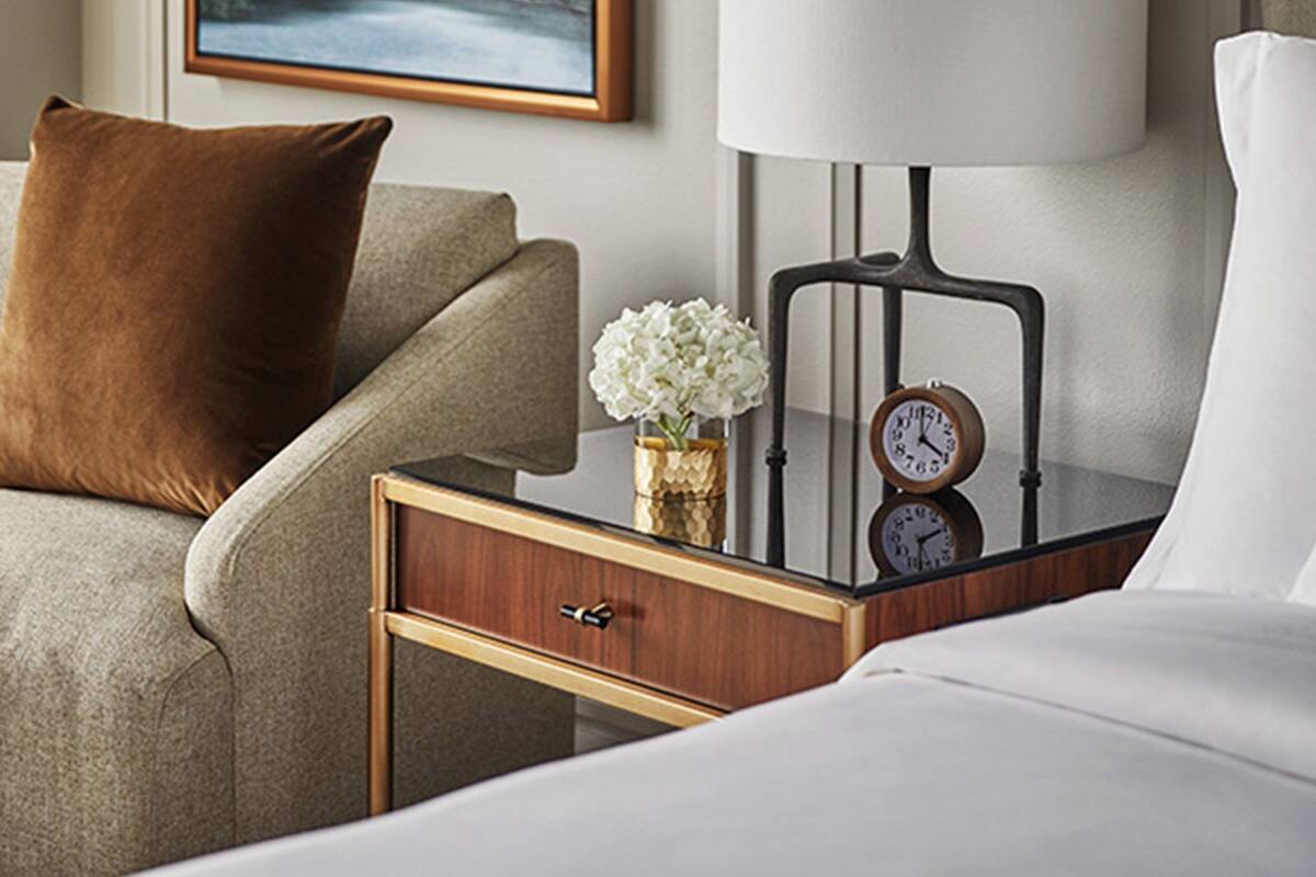 Ein Hotelzimmer mit einem Bett, einem Nachttisch mit einer Uhr, einer Lampe, einem gerahmten Bild an der Wand und einem braunen Kissen auf der Couch.