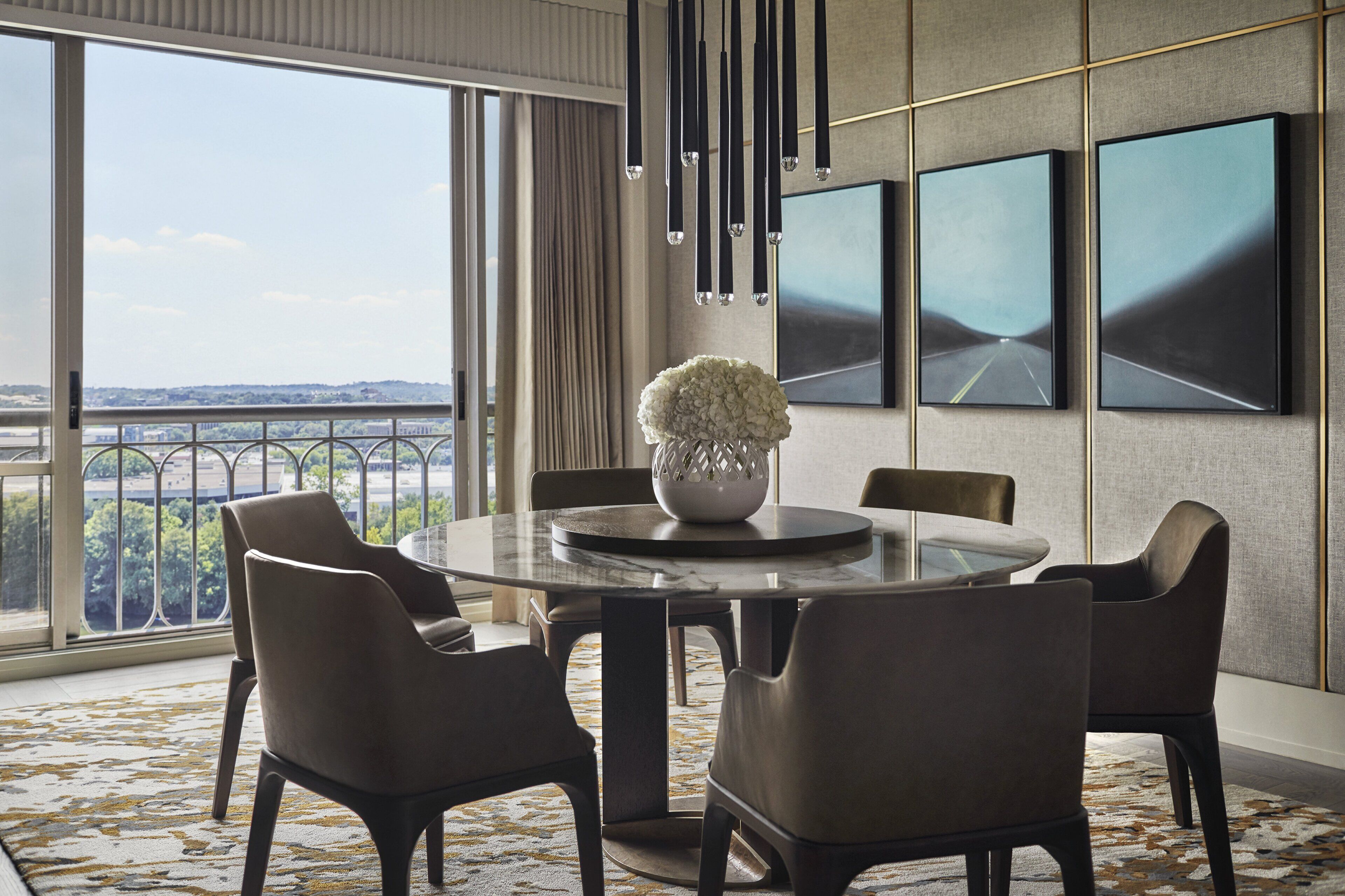A modern dining area with a round table, six chairs, a vase of flowers, and a city view through large windows.