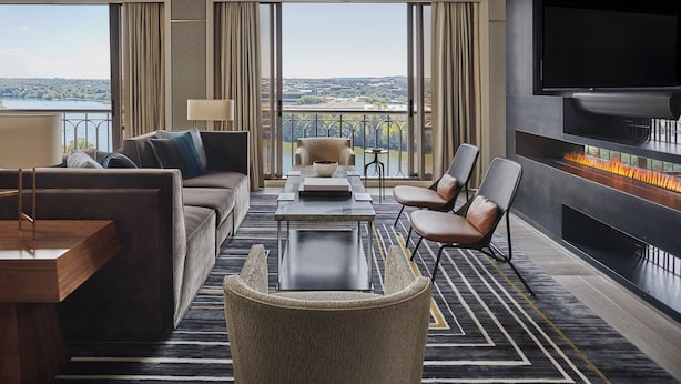 A modern hotel room with a sofa, armchair, and a table with a bowl. A fireplace is visible, and the room has large windows with a view of a cityscape. - Four Seasons Hotel Austin (Austin)