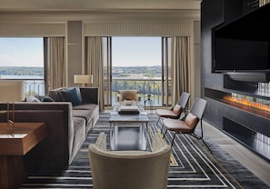 Une chambre d’hôtel moderne avec un canapé, un fauteuil et une table sur laquelle se trouve un bol. On aperçoit une cheminée, et la chambre est dotée de grandes fenêtres offrant une vue sur le paysage urbain.