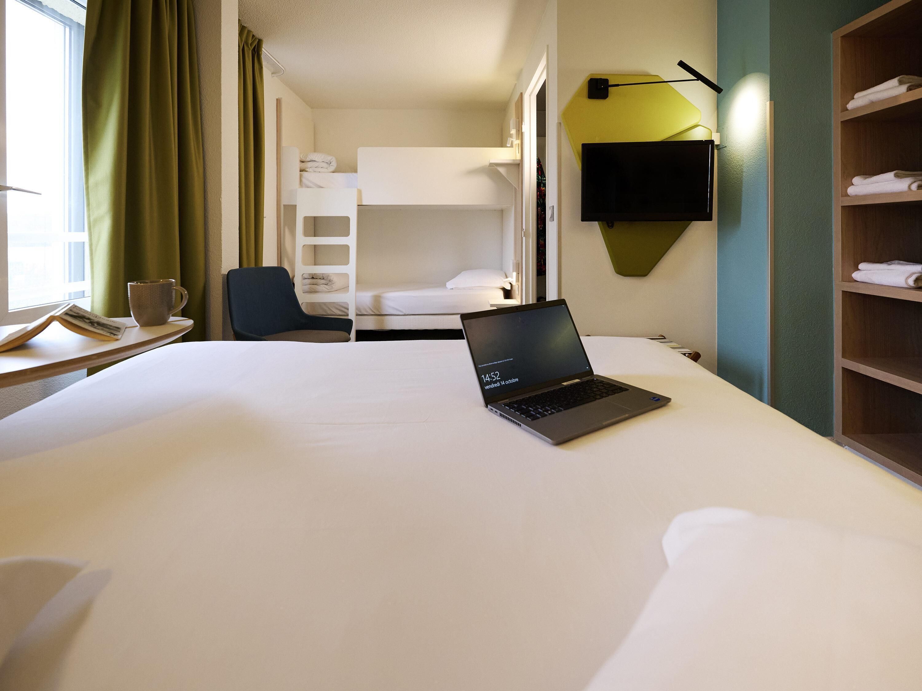 A hotel room with a bed, a laptop, a chair, a TV, and a shelf with towels.