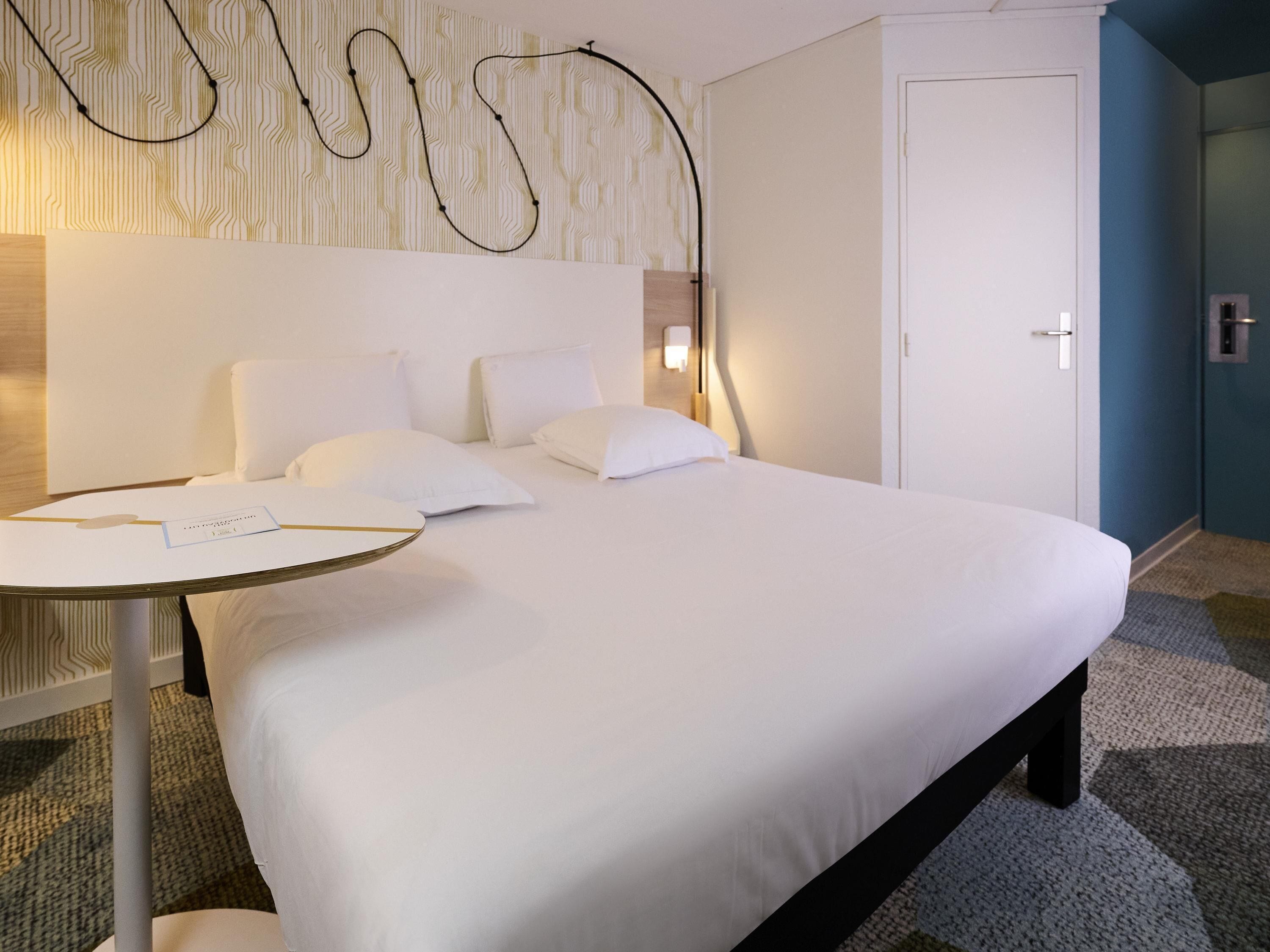 Une chambre d’hôtel avec un lit bien fait, une petite table ronde sur laquelle est posée une carte, une lampe fixée au mur et une porte.