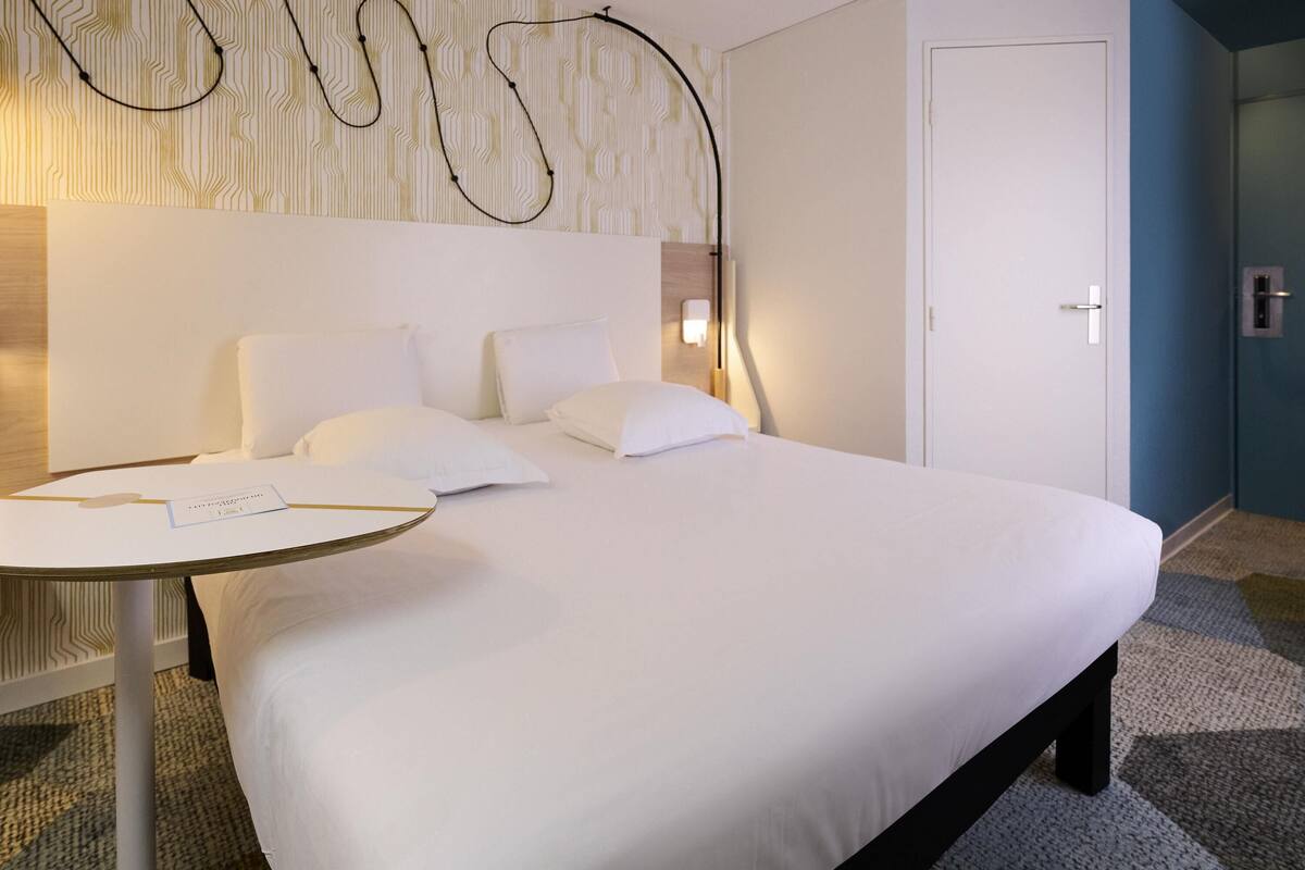 A hotel room with a neatly made bed, a small round table with a card, a wall-mounted light, and a door.