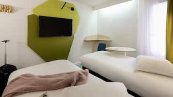 Quarto de hotel com duas camas, uma televisão presa a uma parede verde, uma mesinha e uma janela com cortinas.
