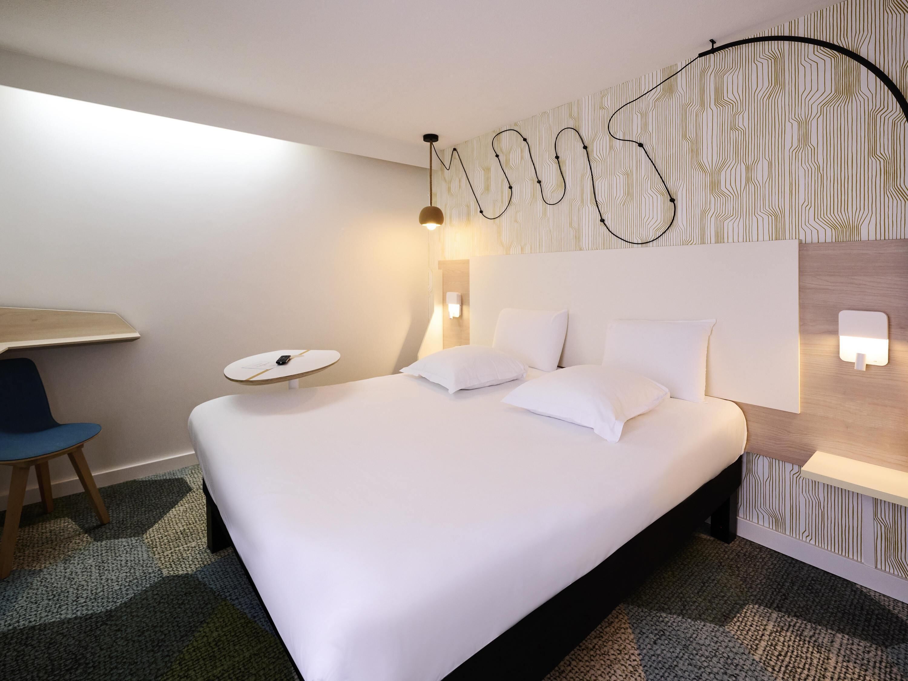 A hotel room with a bed, bedside table, chair, and a wall with a unique pattern.