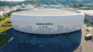 Un estadio grande y moderno con el nombre "ARKEA ARENA" en su fachada, rodeado de un amplio estacionamiento y zonas verdes.