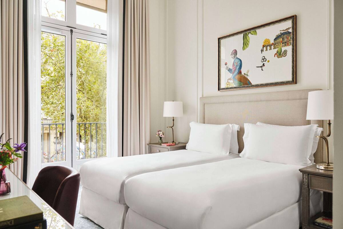 A hotel room with two beds, a window with curtains, a painting on the wall, and a small table with a lamp.
