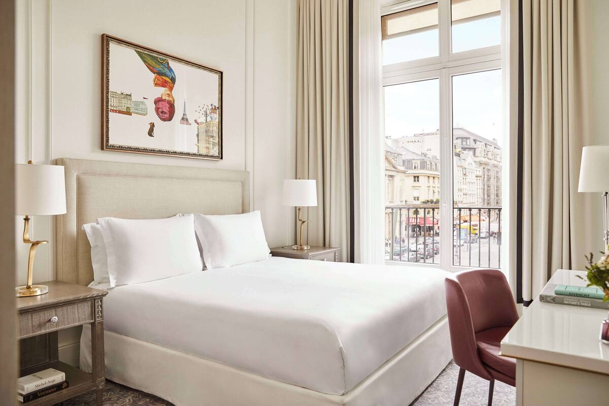 A hotel room with a large bed, two bedside tables with lamps, a chair, a desk with a lamp, a window with a view of buildings, and a framed picture on the wall.