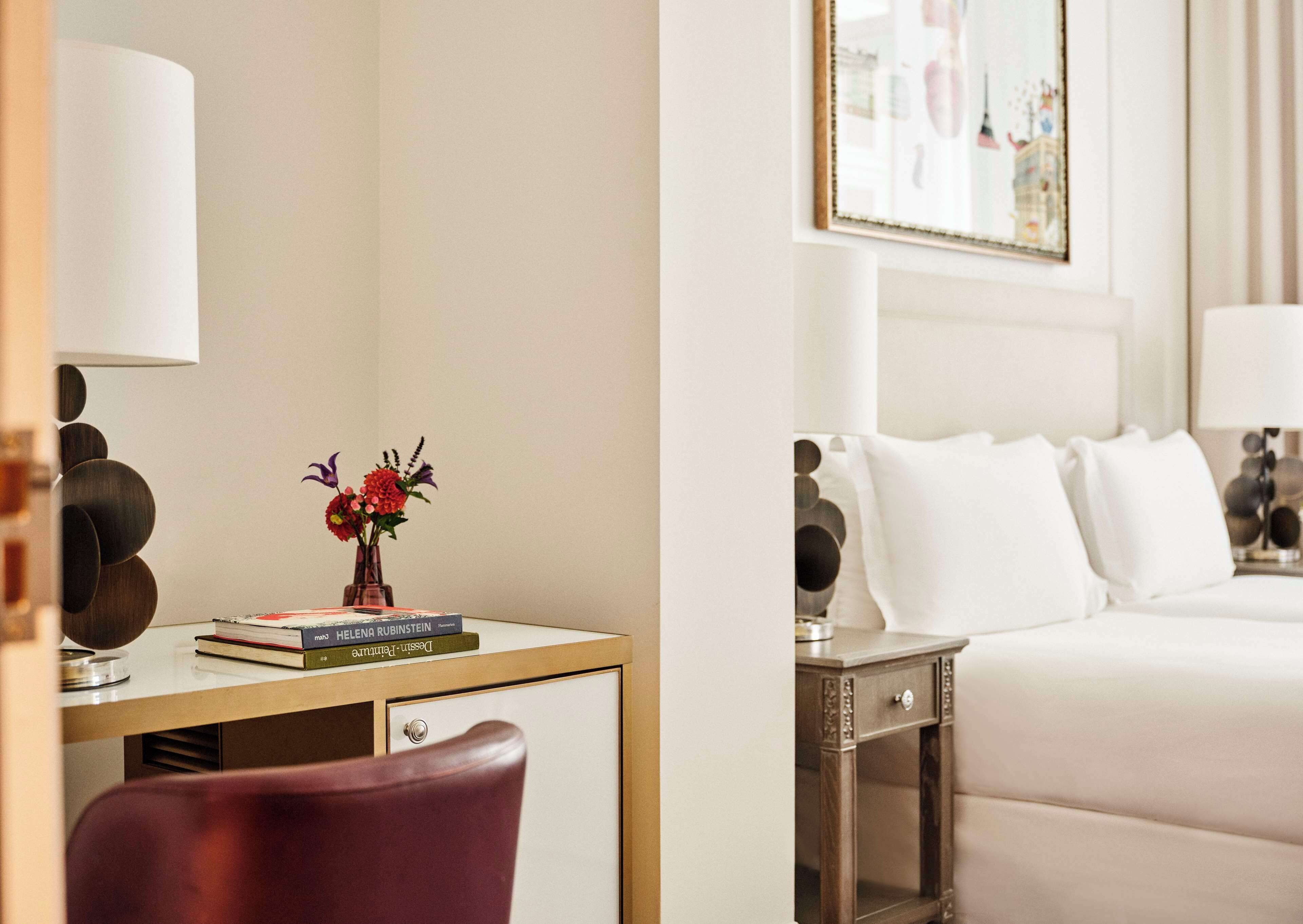 Une chambre d’hôtel avec un lit, une table de chevet, une lampe et un bureau avec des livres et un vase de fleurs.