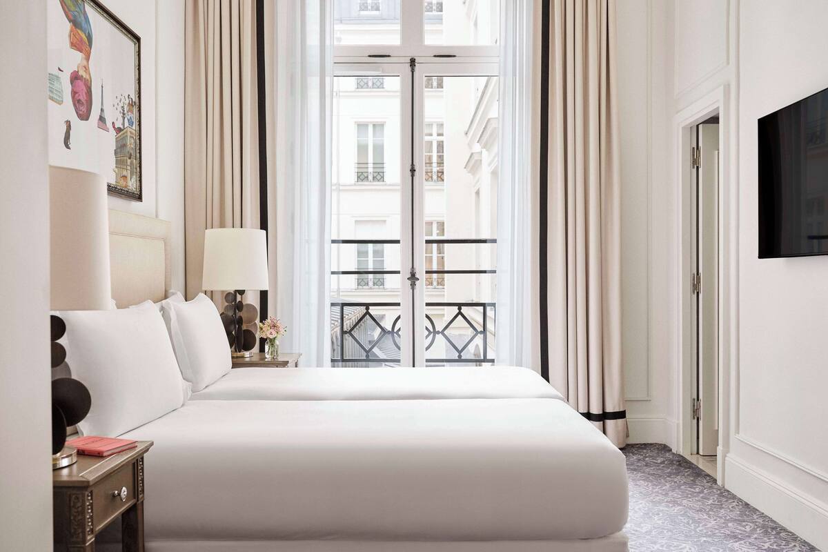 A hotel room with a large bed, a bedside table with a lamp, a TV on the wall, and a view of the city through a balcony door.