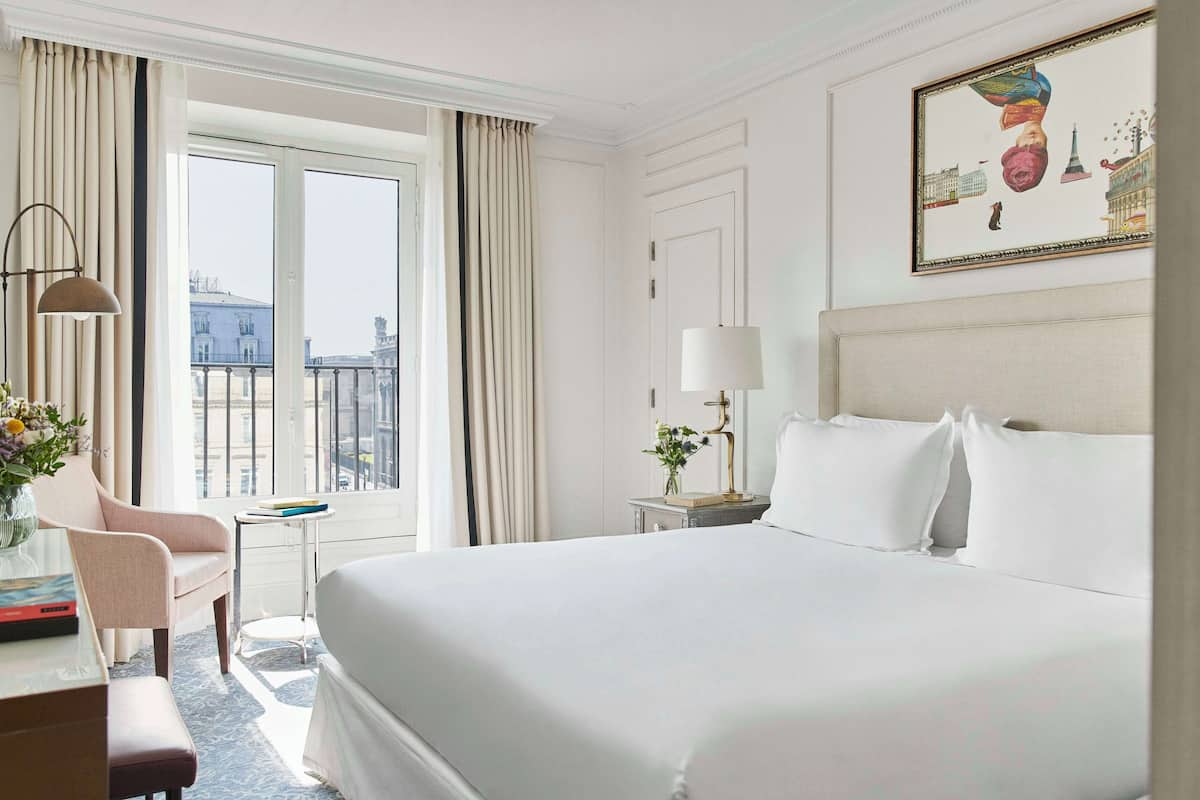 Ein Hotelzimmer mit einem großen Bett, einem Schreibtisch mit Stuhl, einem kleinen Tisch mit Lampe und einem Fenster mit Vorhängen.