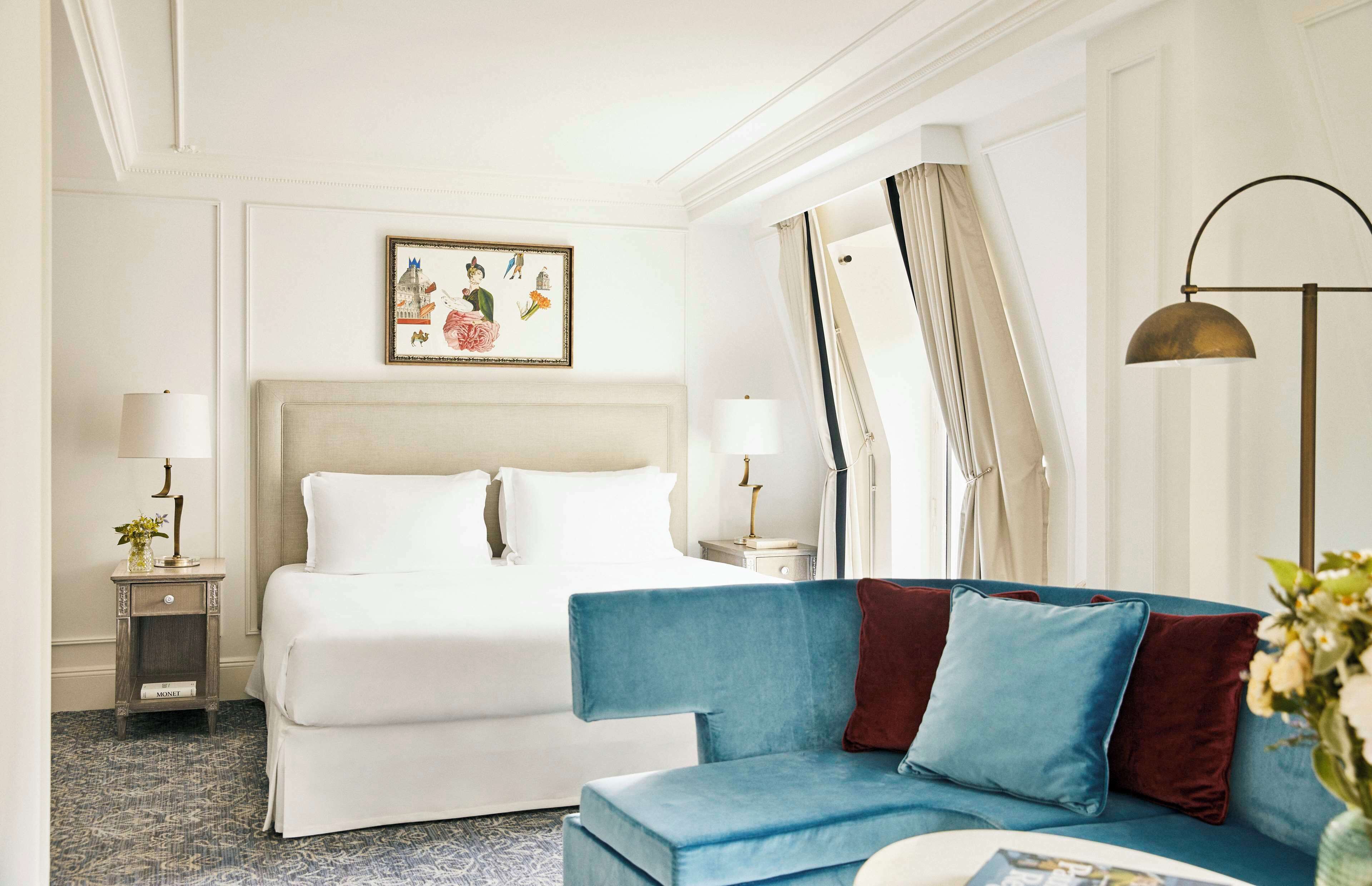 A hotel room with a bed, a blue sofa, a nightstand with a lamp, and a painting on the wall.