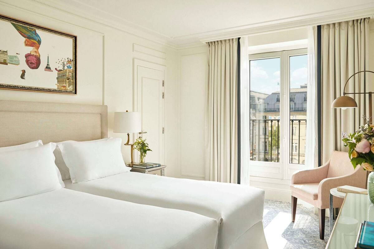 A hotel room with two beds, a desk with a lamp, a chair, a painting, and a balcony with curtains.