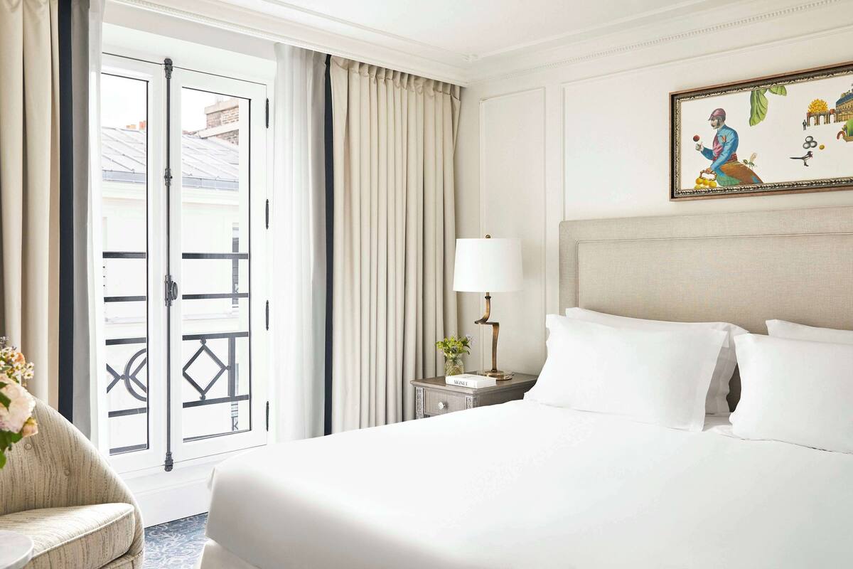A hotel room with a bed, a nightstand with a lamp, a chair, a vase with flowers, and a painting on the wall.