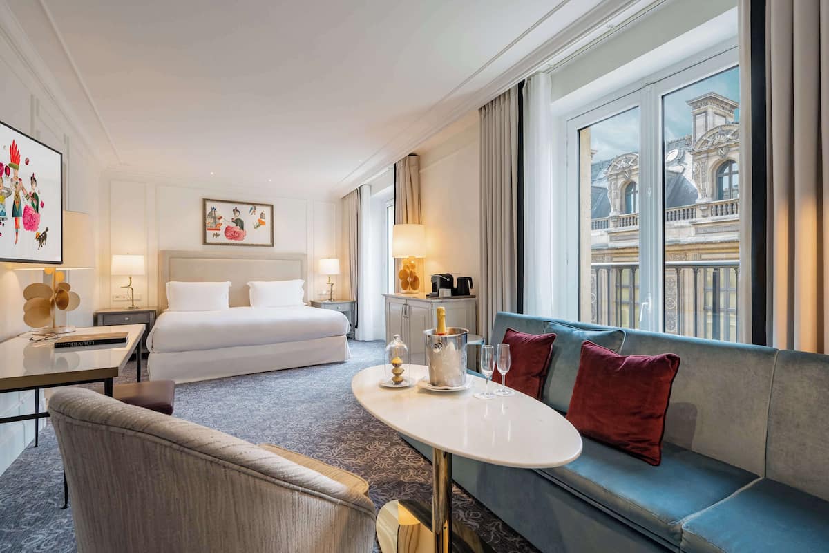 Ein Hotelzimmer mit Bett, einer Sitzecke, einem Tisch mit einer Champagnerflasche und Blick auf ein Gebäude durch das Fenster.