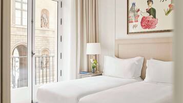 A hotel room with two beds, a large window with a view of a building, a painting on the wall, and a nightstand with a lamp and a vase of flowers.