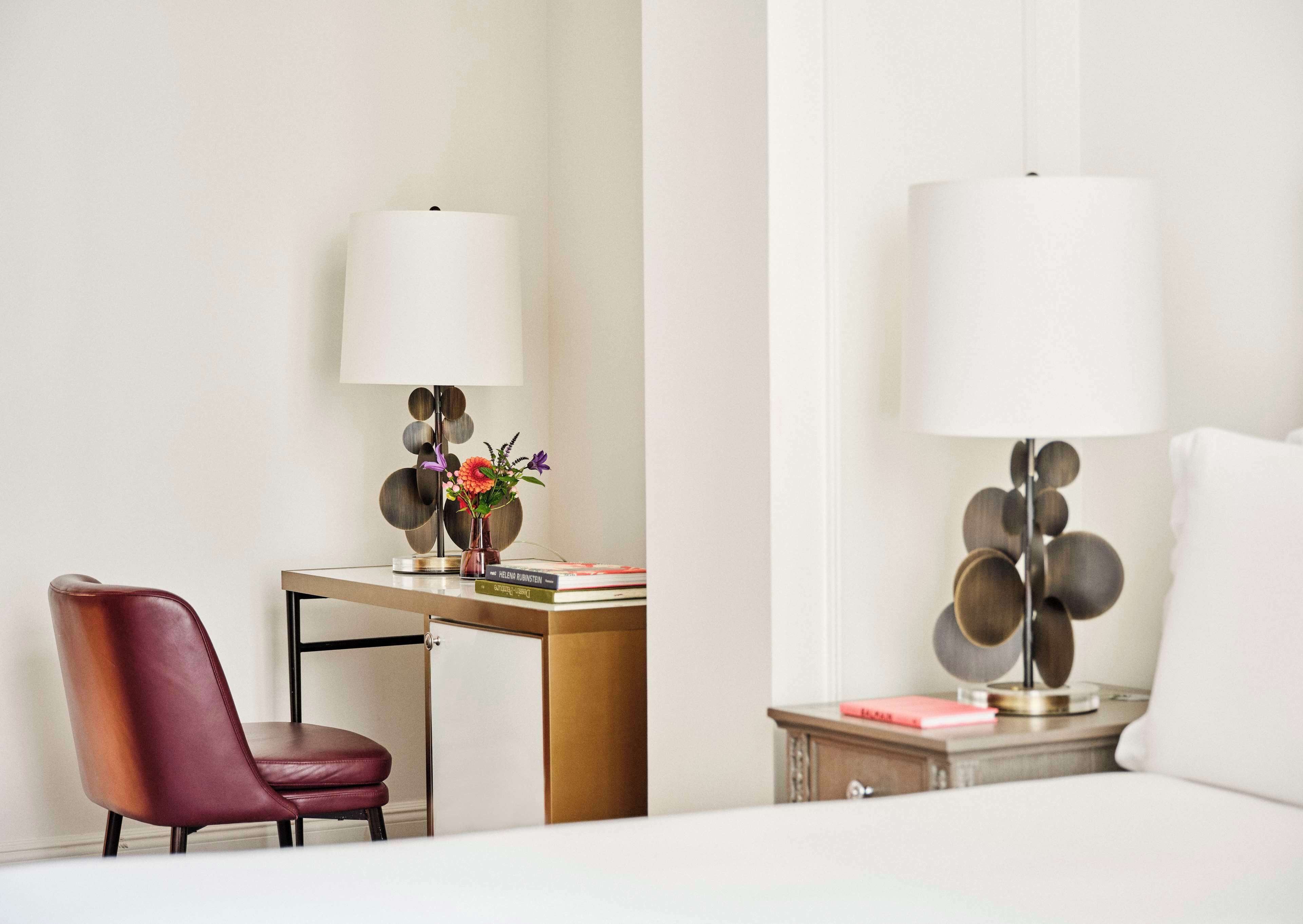 A modern hotel room with a desk, a chair, a bedside table with a lamp, and a bed.