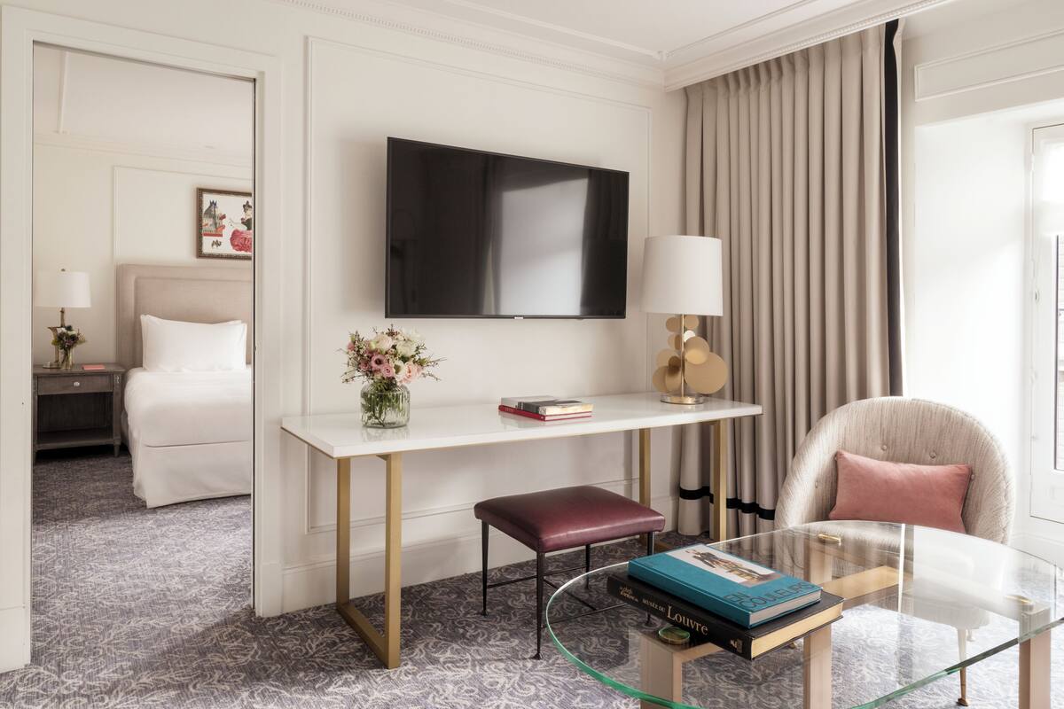 A hotel room with a bed, a desk with a flat-screen TV, a glass coffee table with books, a chair, and a window with curtains.
