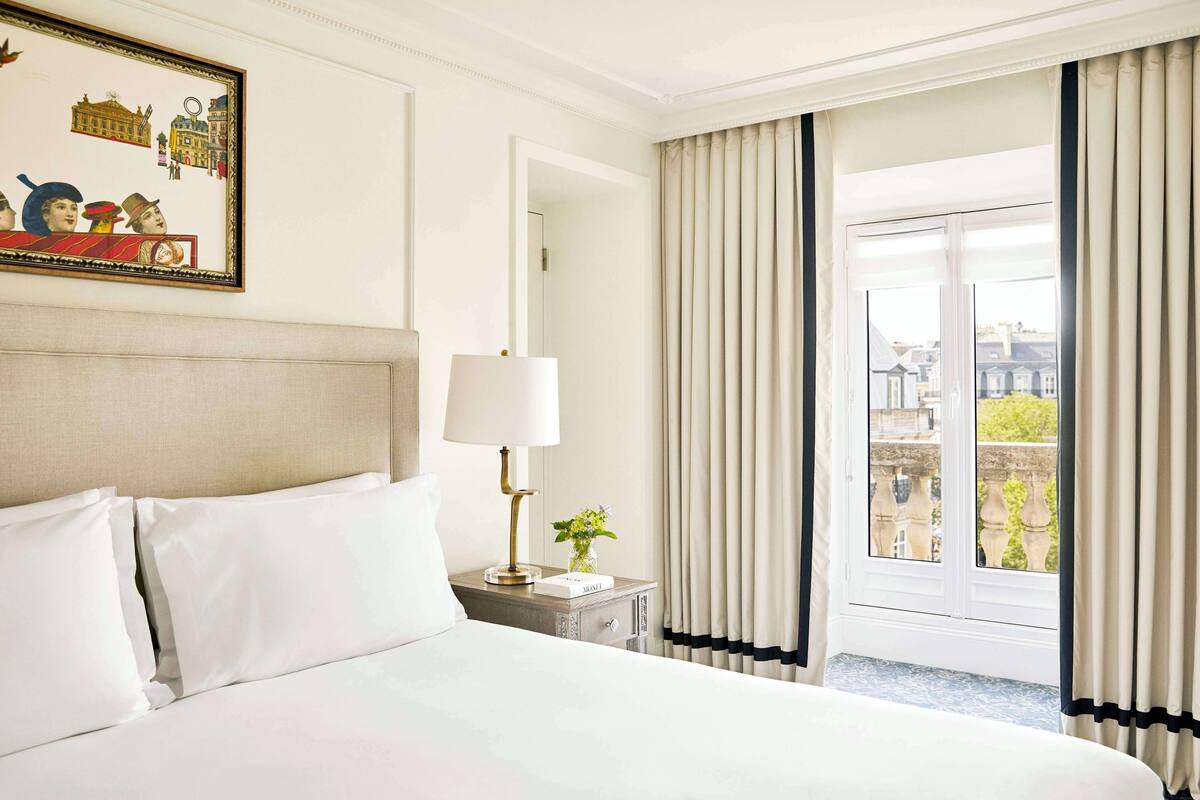 A hotel room with a neatly made bed, a bedside lamp, a framed picture on the wall, and a window with curtains.