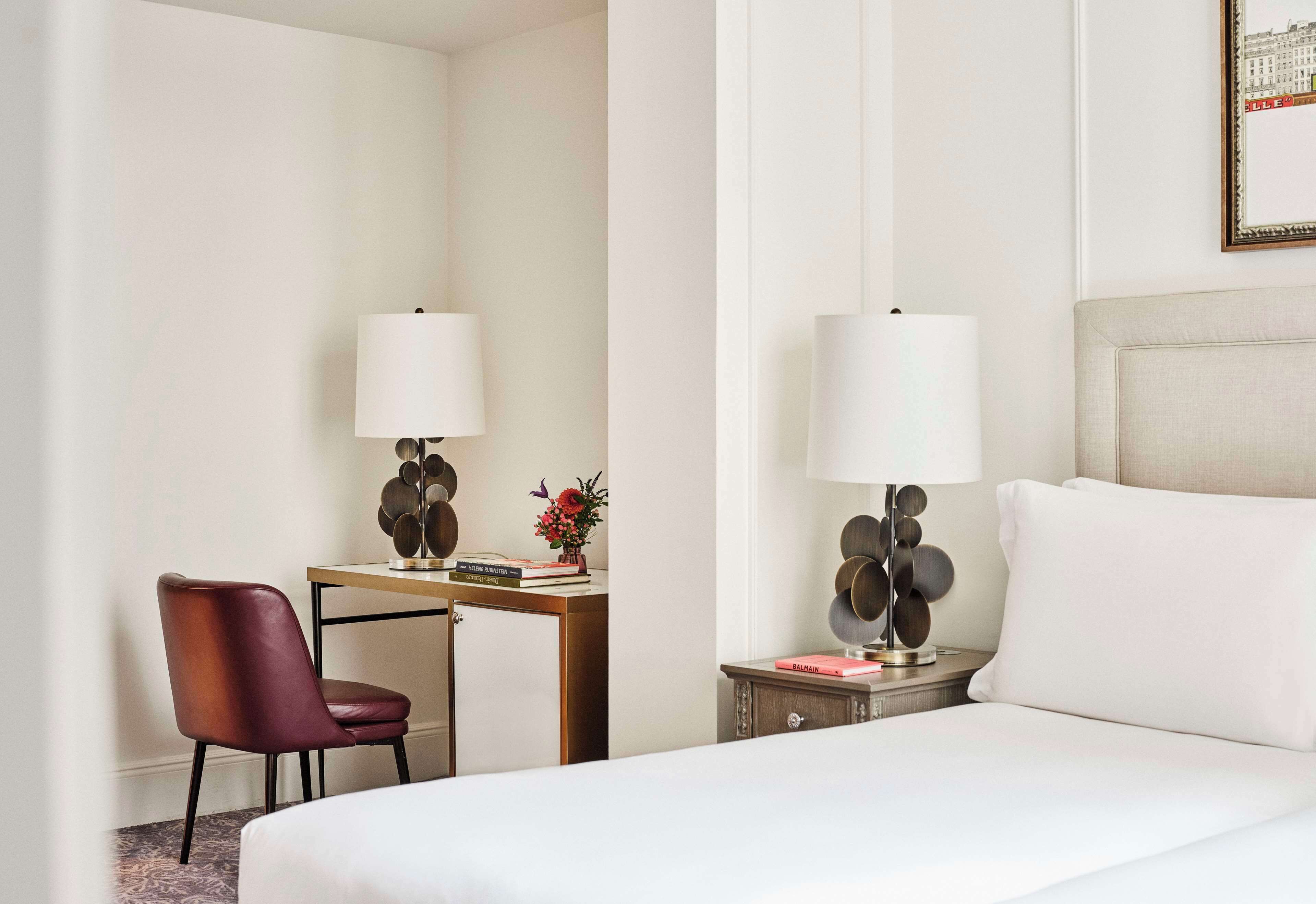 A hotel room with a bed, a desk with a chair, a lamp, and a small table with a vase of flowers.