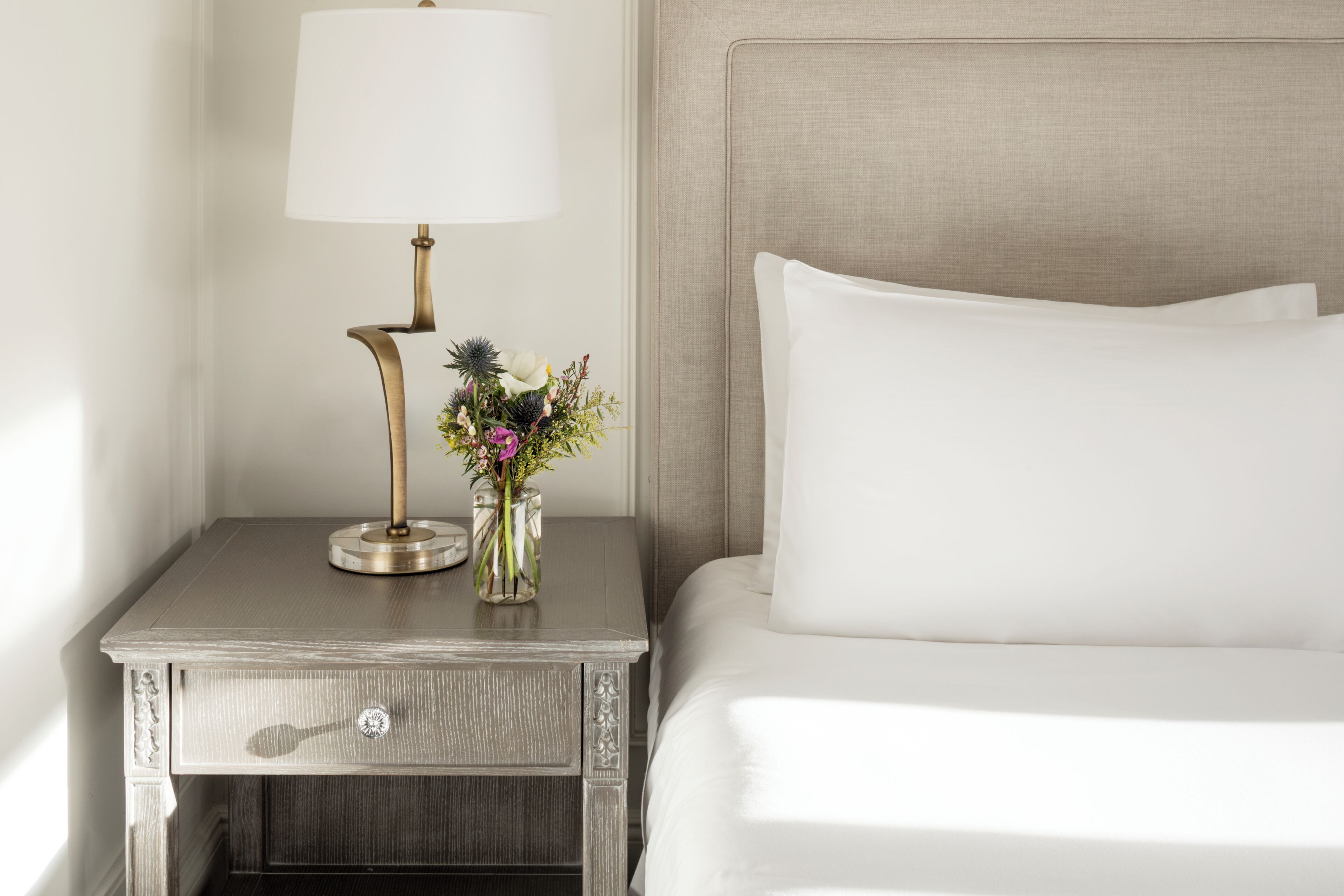 A neatly made bed with a headboard, a bedside table with a lamp, and a vase with flowers.