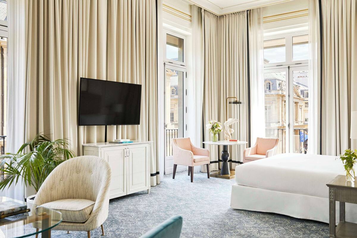 A hotel room with a large bed, a TV, a desk with a chair, a small table with a vase, and a potted plant.