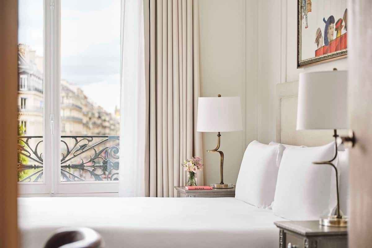 Ein Hotelzimmer mit großem Fenster, einem Bett mit weißer Bettwäsche, einem Nachttisch mit Lampe und einem Gemälde an der Wand.