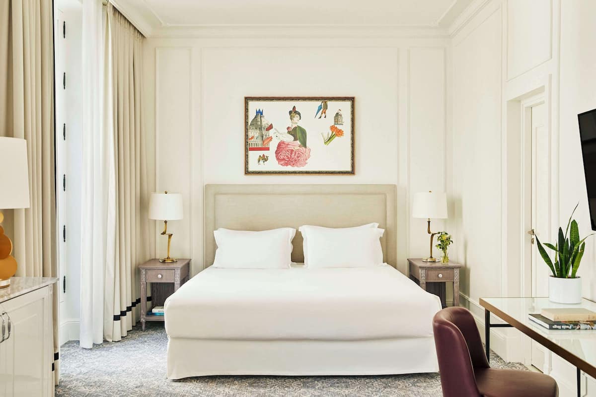 Ein Hotelzimmer mit einem großen Bett, zwei Nachttischen mit Lampen, einem Schreibtisch mit Stuhl, einem gerahmten Bild an der Wand und einer Zimmerpflanze.