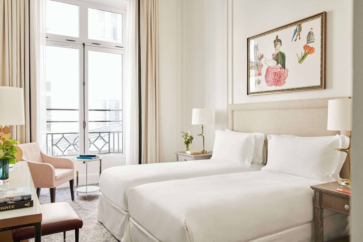 A hotel room with two beds, a desk, a chair, a window with a view, and a painting on the wall.