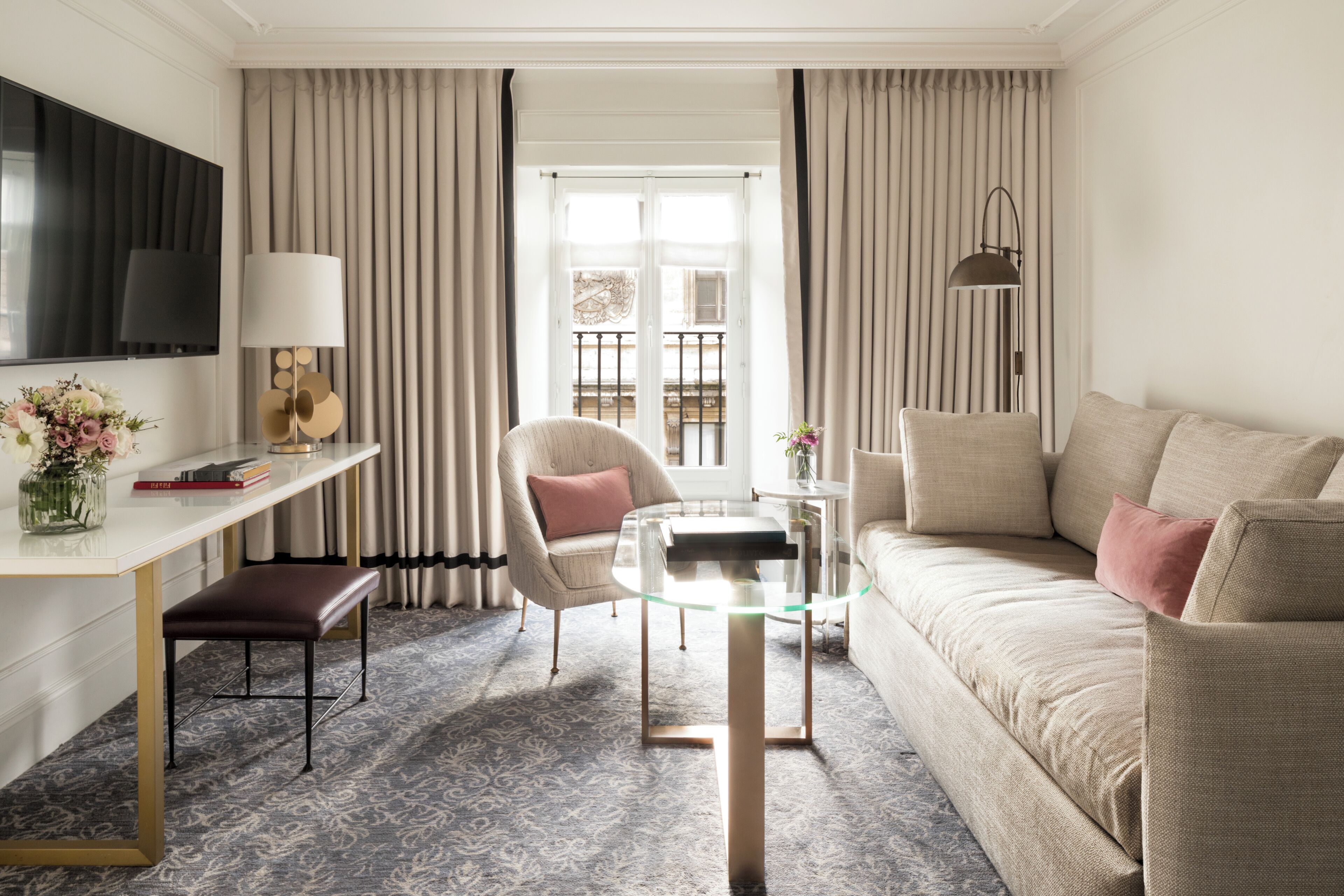 A hotel room with a sofa, a glass coffee table, a chair, a desk, a TV, and a window with curtains.
